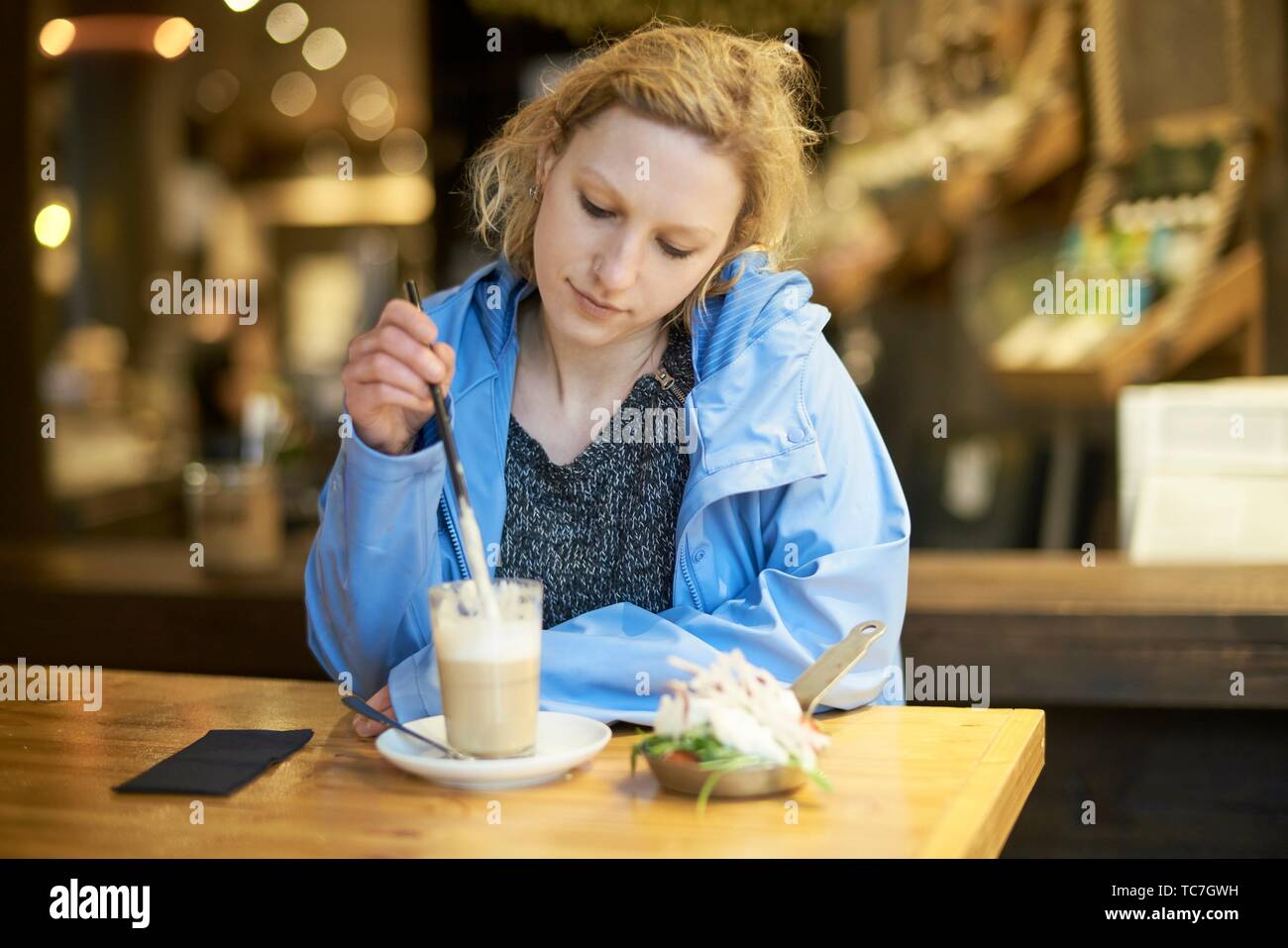 Young thoughtful woman stirring latte macchiato with drinking straw, in cafe, taking a break alone, thinking, in Munich, Germany. Stock Photo