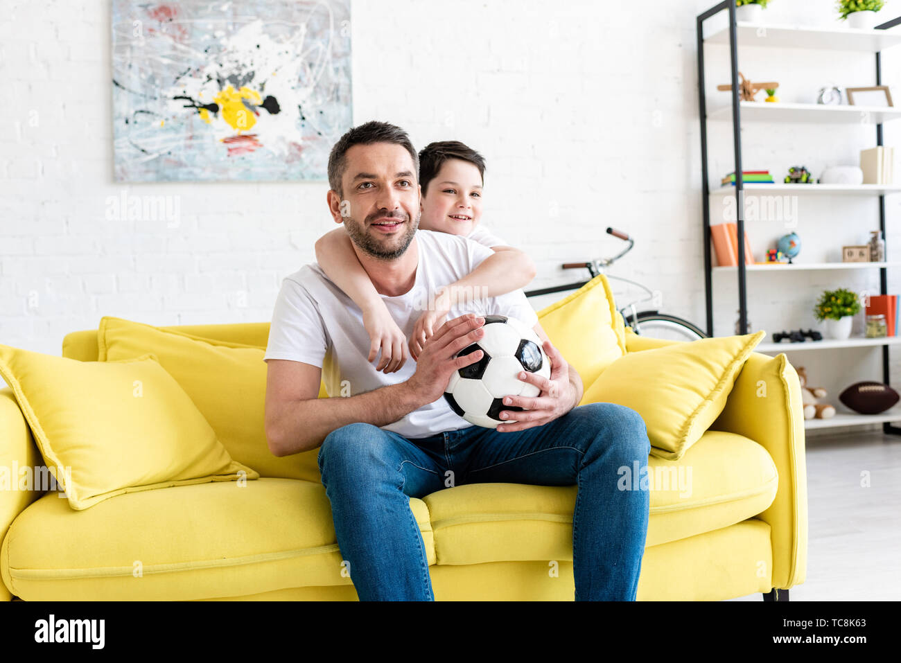 Father And Son Watching Sports Match On Couch With Soccer Ball At