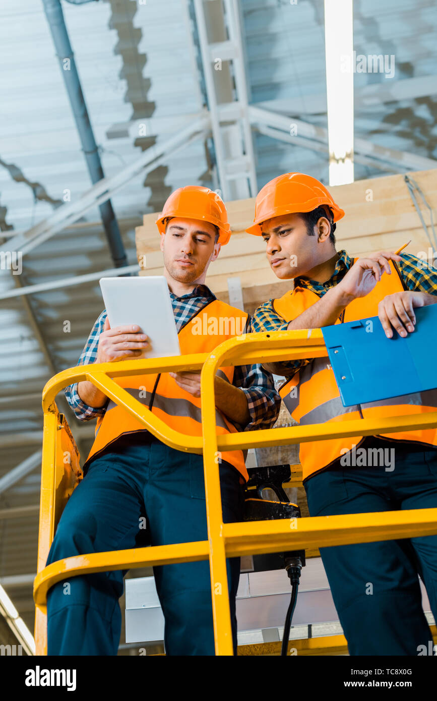 serious multicultural warehouse workers using digital tablet while standing on scissor lift Stock Photo