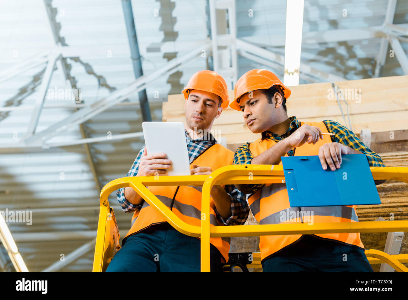 thoughtful multicultural workers using digital tablet while standing on scissor lift in warehouse Stock Photo