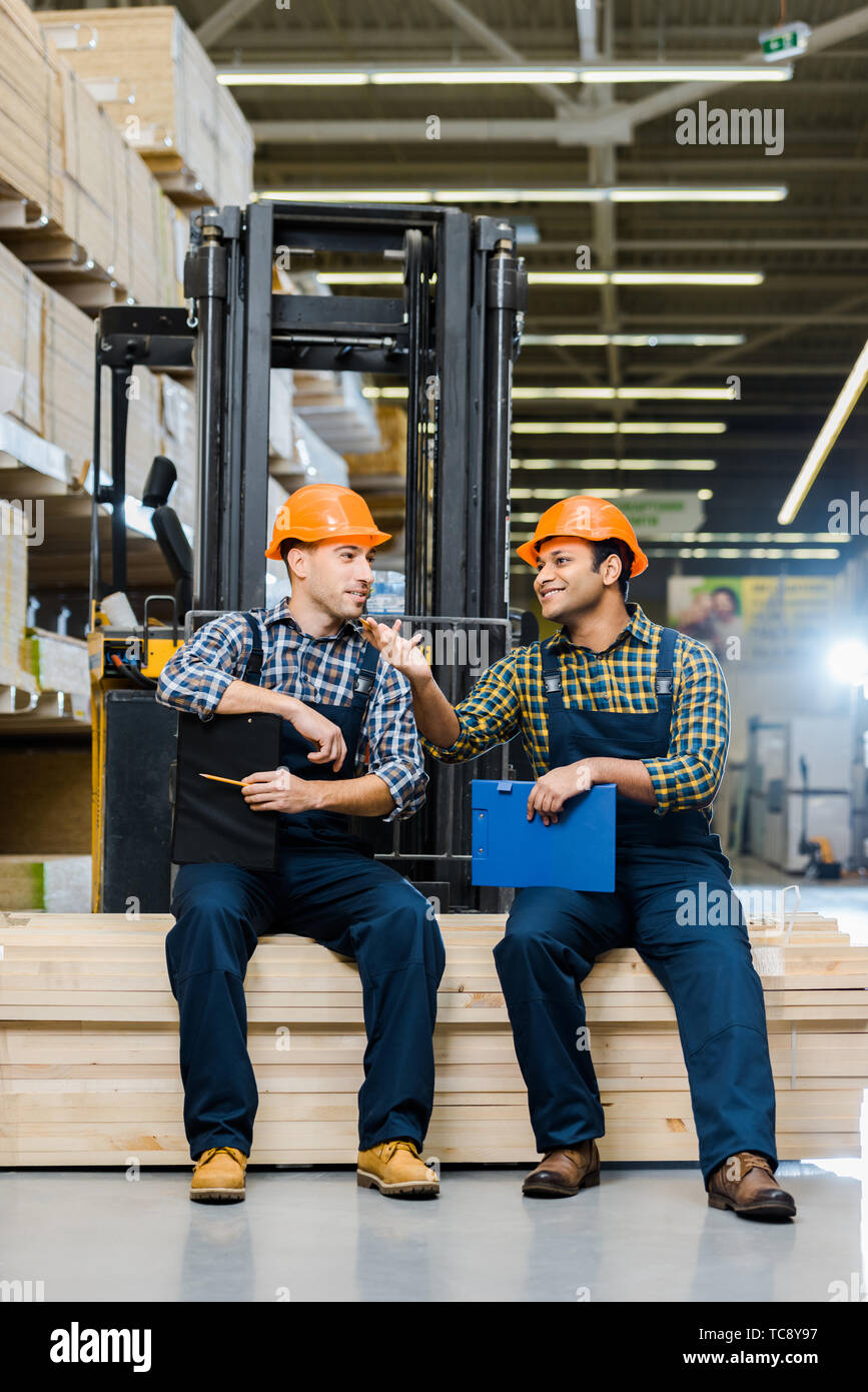two smiling multicultural workers with clipboards talking while sitting ...