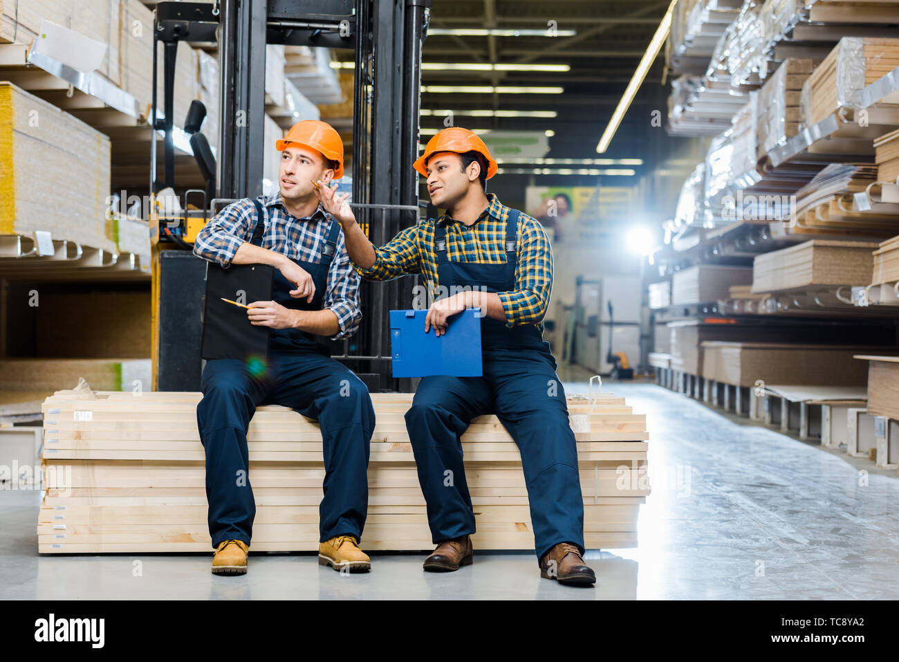 two multicultural workers talking while sitting on plywood in warehouse Stock Photo
