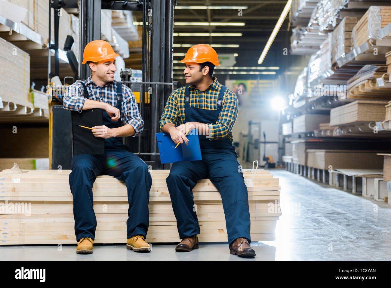 two multicultural workers smiling and talking while sitting on plywood ...