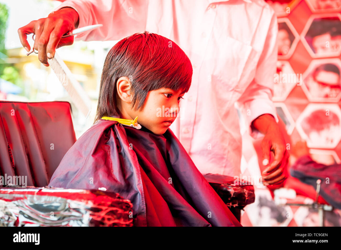 A shy little Cambodian boy get his hair cut in a typical outdoor barbershop in Phnom Penh, Cambodia. Stock Photo