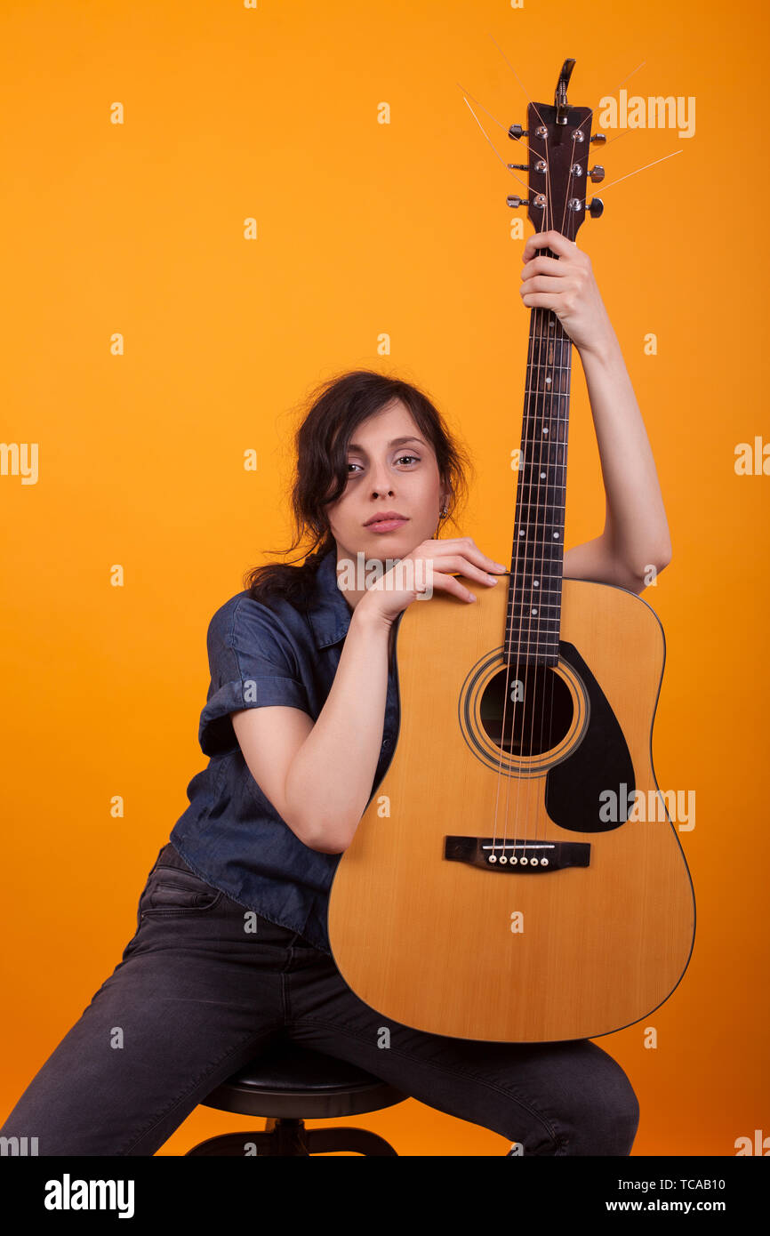 Portrait of beautiful young singer holding her acustic guitar in studio over yellow background. Pretty caucasian woman with musical instrument. Stock Photo