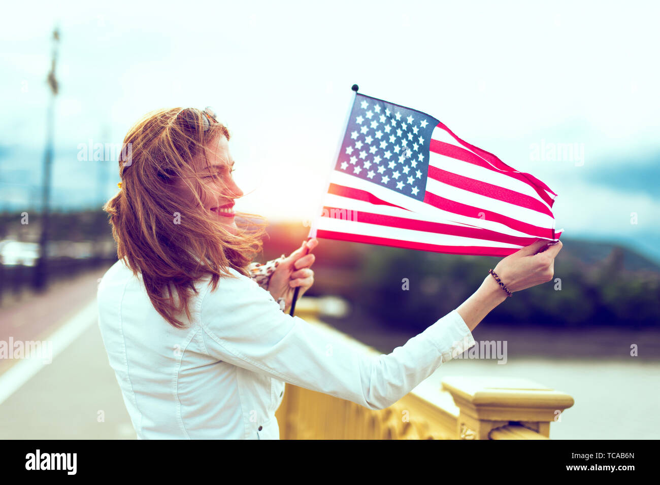 Happy young patriot urban woman with toothy smile stretching USA flag, profile view, with sunlight Stock Photo