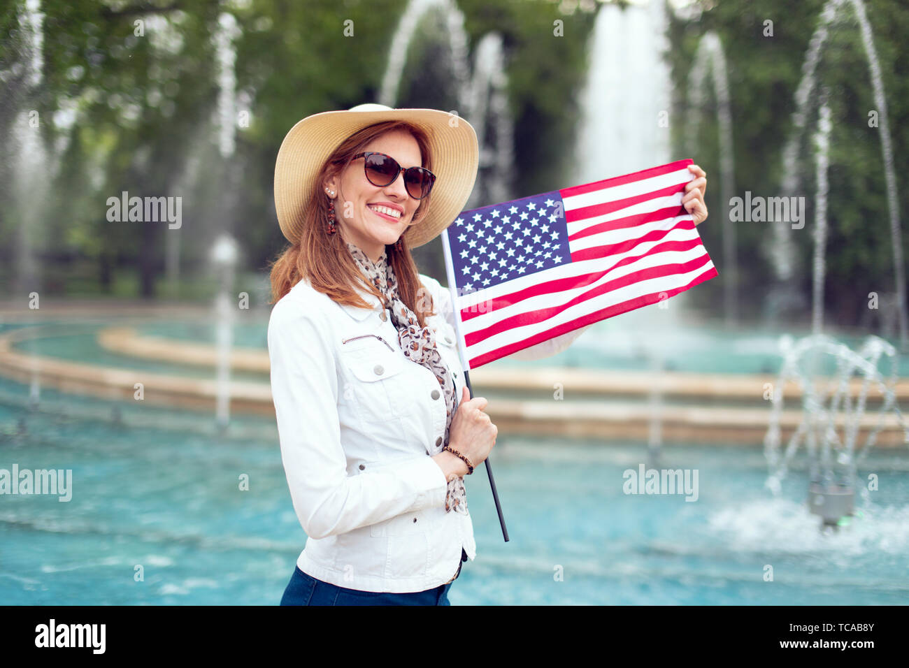 Young fashionable excited woman holding USA flag in park at fountain, Independence day, Fourth of July Stock Photo