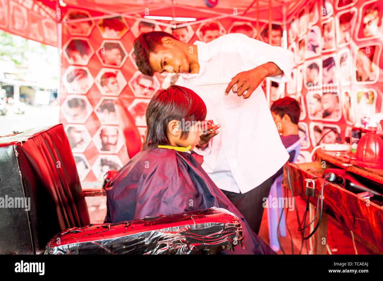 A shy little Cambodian boy get his hair cut in a typical outdoor barbershop in Phnom Penh, Cambodia. Stock Photo