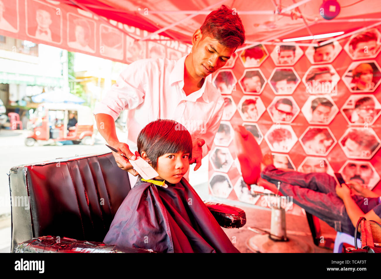 A shy little Cambodian boy get his hair cut in a typical outdoor barbershop in Phnom Penh, Cambodia. Stock Photo