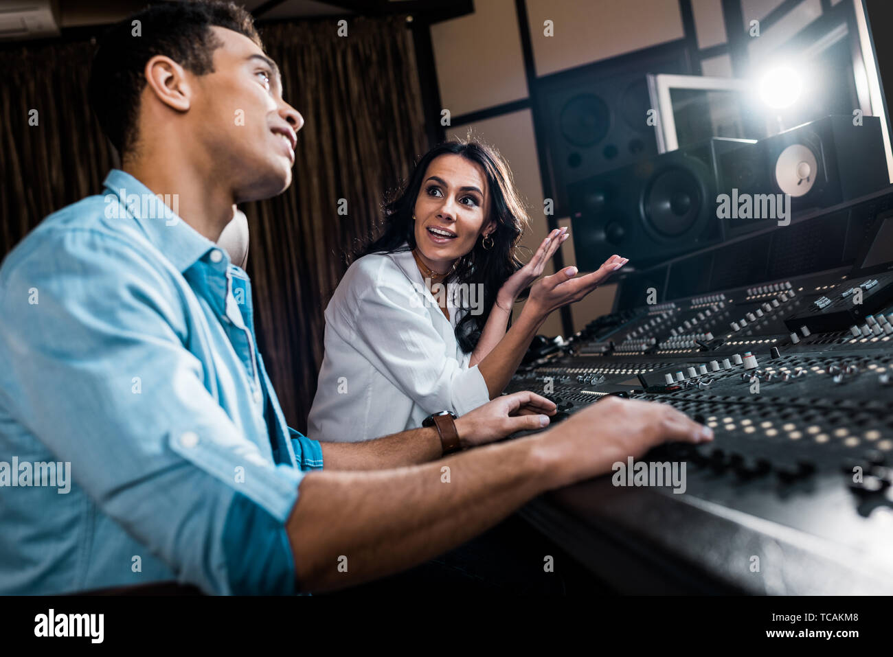 selective focus of pretty sound producer gesturing near mixed raced colleague working at mixing console Stock Photo