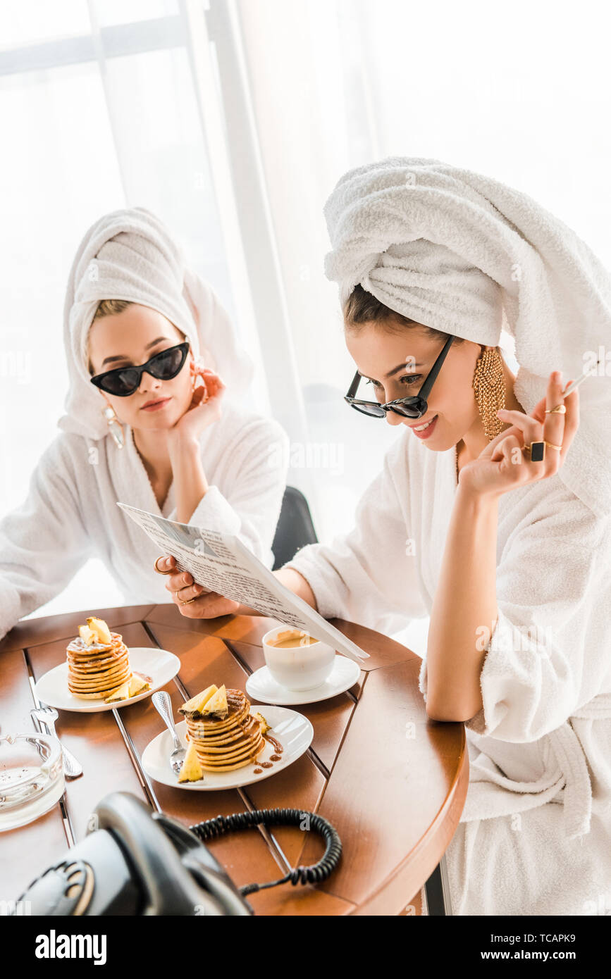 stylish smiling woman in bathrobe, sunglasses and jewelry with towel on head smoking cigarette and reading newspaper with friend at morning Stock Photo
