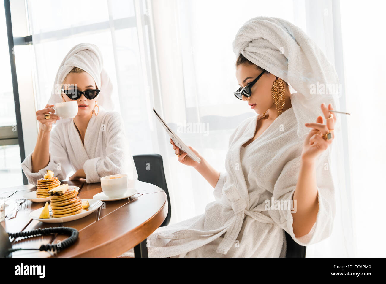 stylish women in bathrobes, sunglasses and jewelry with towels on heads smoking cigarette and reading newspaper at morning Stock Photo