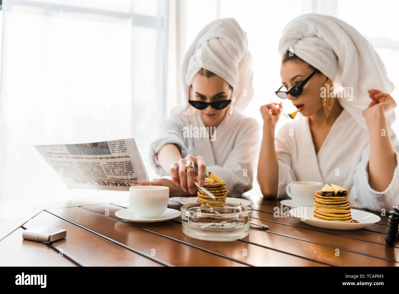selective focus of stylish women in bathrobes, sunglasses and jewelry with towels on heads smoking cigarette and reading newspaper while eating pancak Stock Photo
