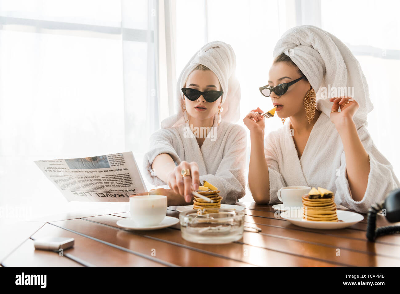 stylish women in bathrobes, sunglasses and jewelry with towels on heads smoking cigarette and reading newspaper while eating pancakes Stock Photo