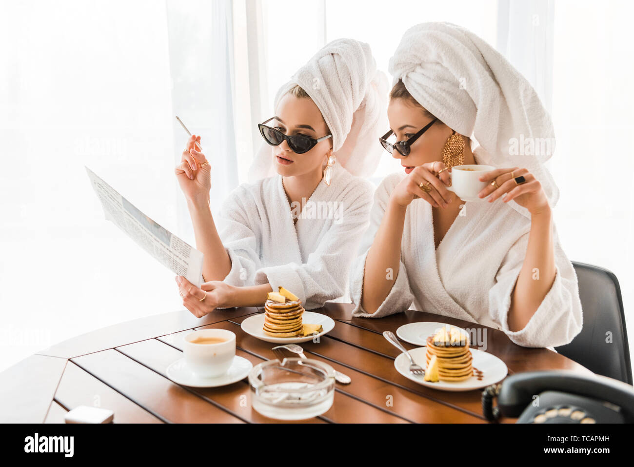 stylish women in bathrobes, sunglasses and jewelry with towels on heads smoking cigarette and reading newspaper while having breakfast Stock Photo