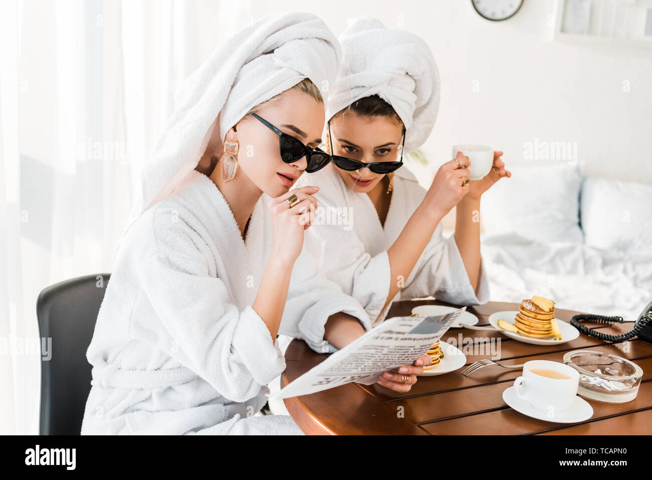 stylish women in bathrobes, sunglasses and jewelry with towels on heads smoking and reading newspaper while having breakfast Stock Photo