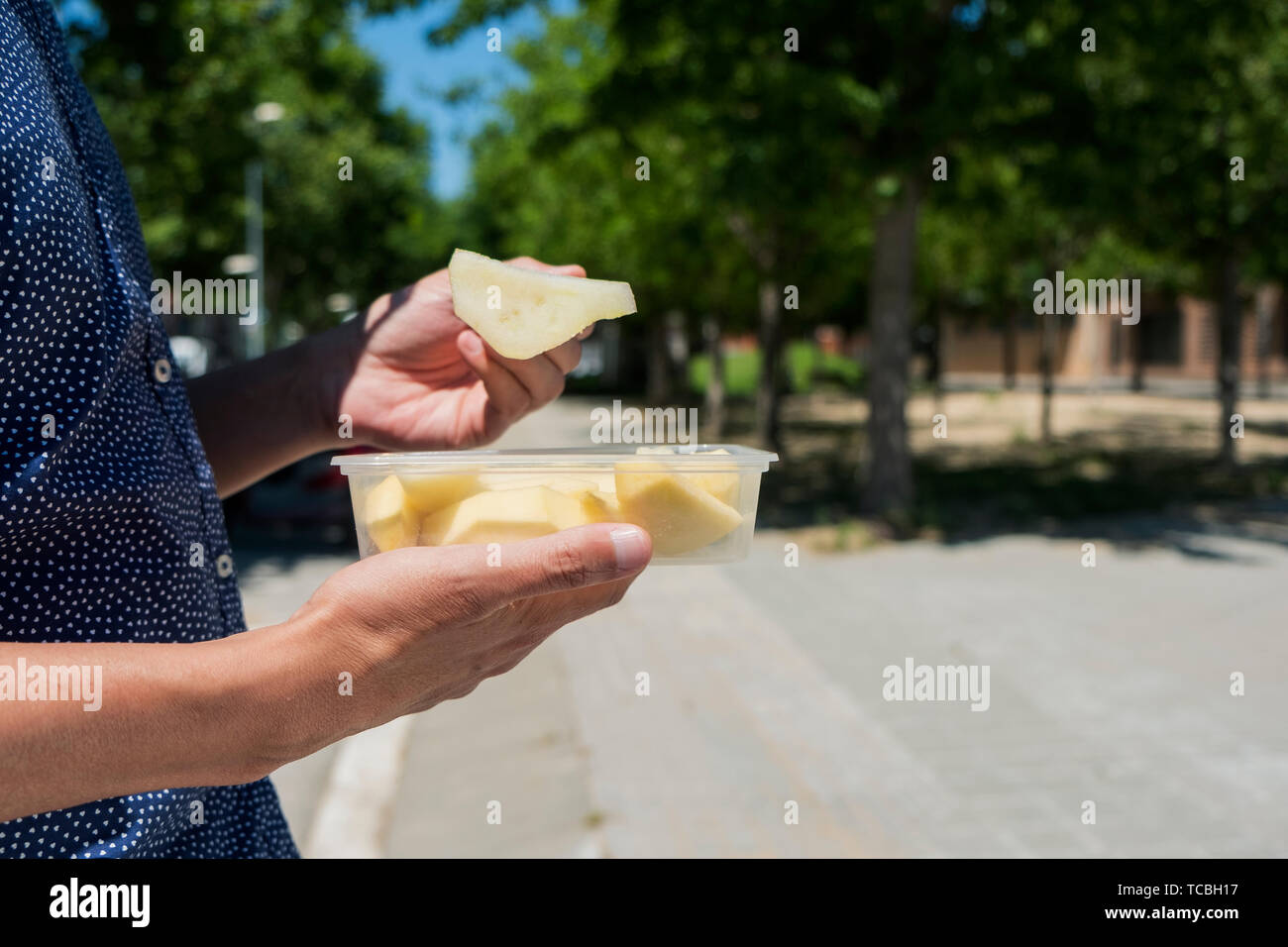 closeup of a young caucasian man outdoors, wearing a blue short-sleeved shirt, eating a salad fruit, made with pear, apple and peach, from a plastic c Stock Photo