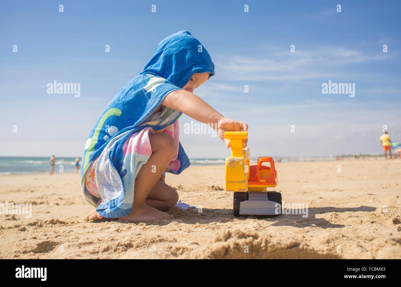Baby boy playing on sand at the beach with excavator toy. He is wearing hooded poncho towel. Stock Photo