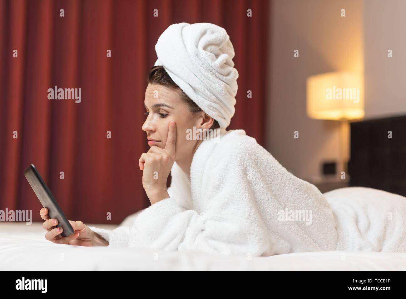 Young woman in bathrobe lying in hotel room using mobile phone, relaxed