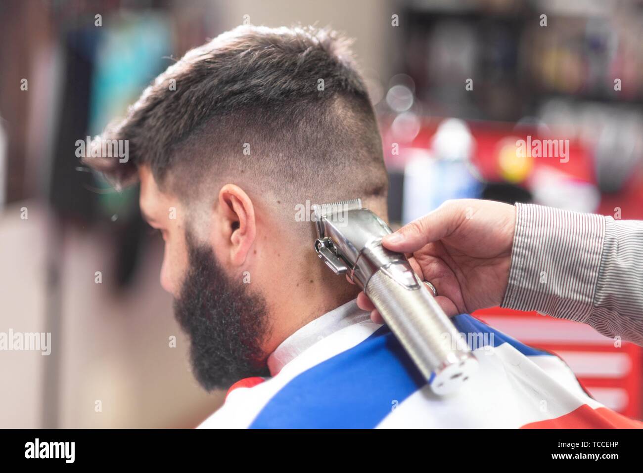 Handsome bearded man, getting haircut by barber, with electric trimmer at barbershop. Stock Photo