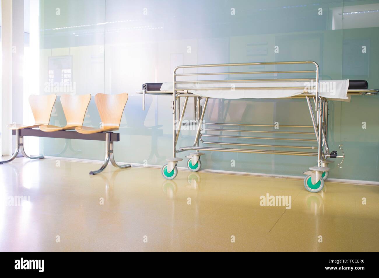 Interior of healthcare center hallway. Modern and gleaming environment with stretcher and wooden waiting chairs. Stock Photo