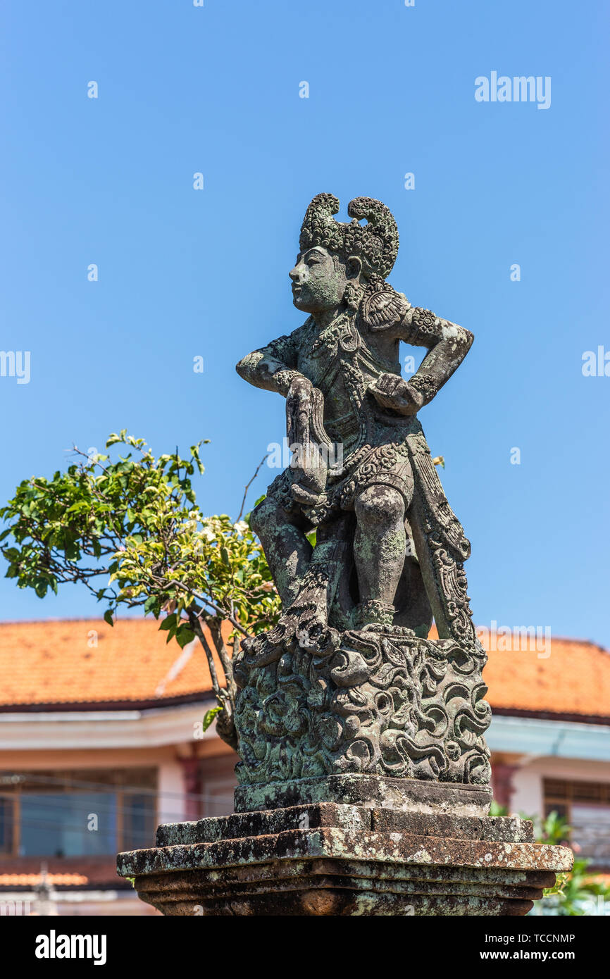 Klungkung, Bali, Indonesia - February 26, 2019: Closeup of Stone statue of defiant King covered in black mold against blue sky. Some green foliage and Stock Photo