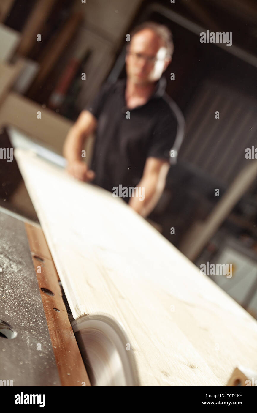 Carpenter, joiner or woodworking working in a workshop with long planks of wood guiding them through a saw in the foreground Stock Photo