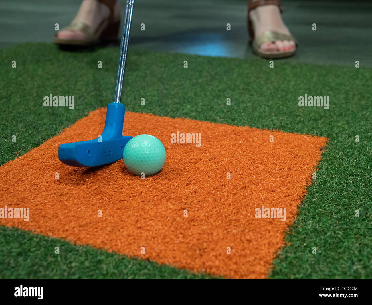 Blue putter on turf next to green golf ball on miniature golf course with woman hitting Stock Photo