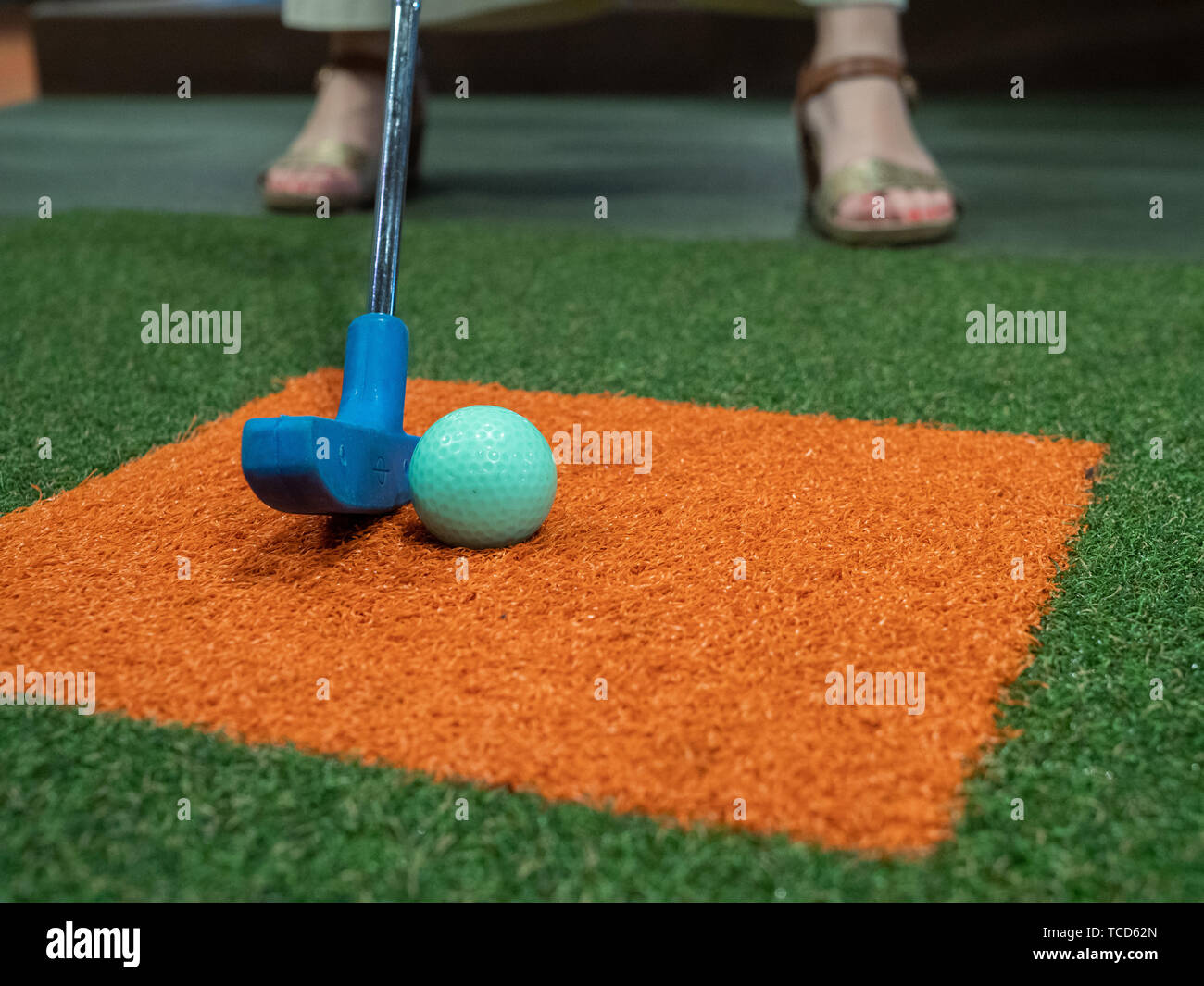 Blue putter on turf lined up next to green golf ball on miniature golf course with woman playing Stock Photo