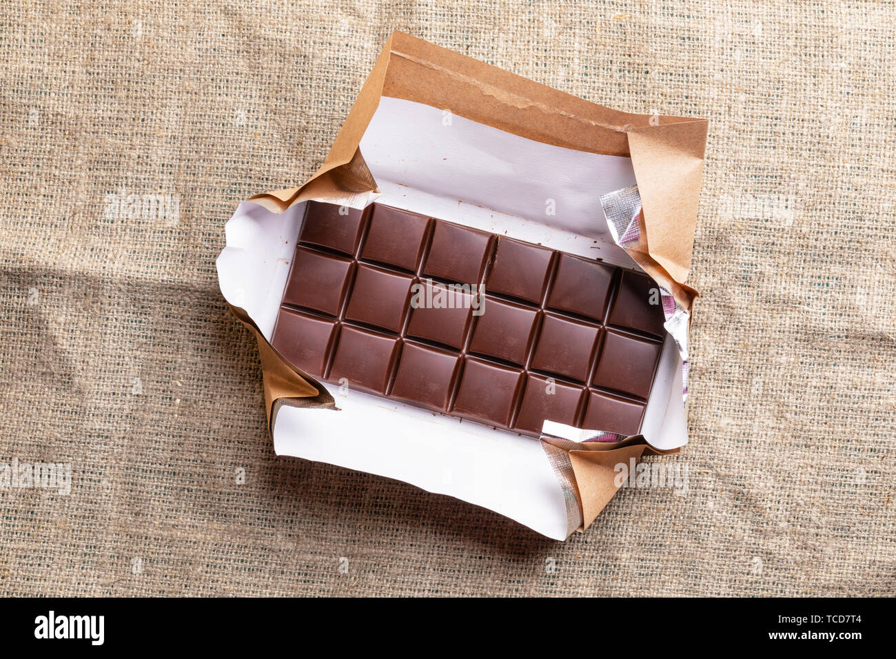 Whole chocolate bar in opened paper wrapper is lying on burlap.  Stock Photo
