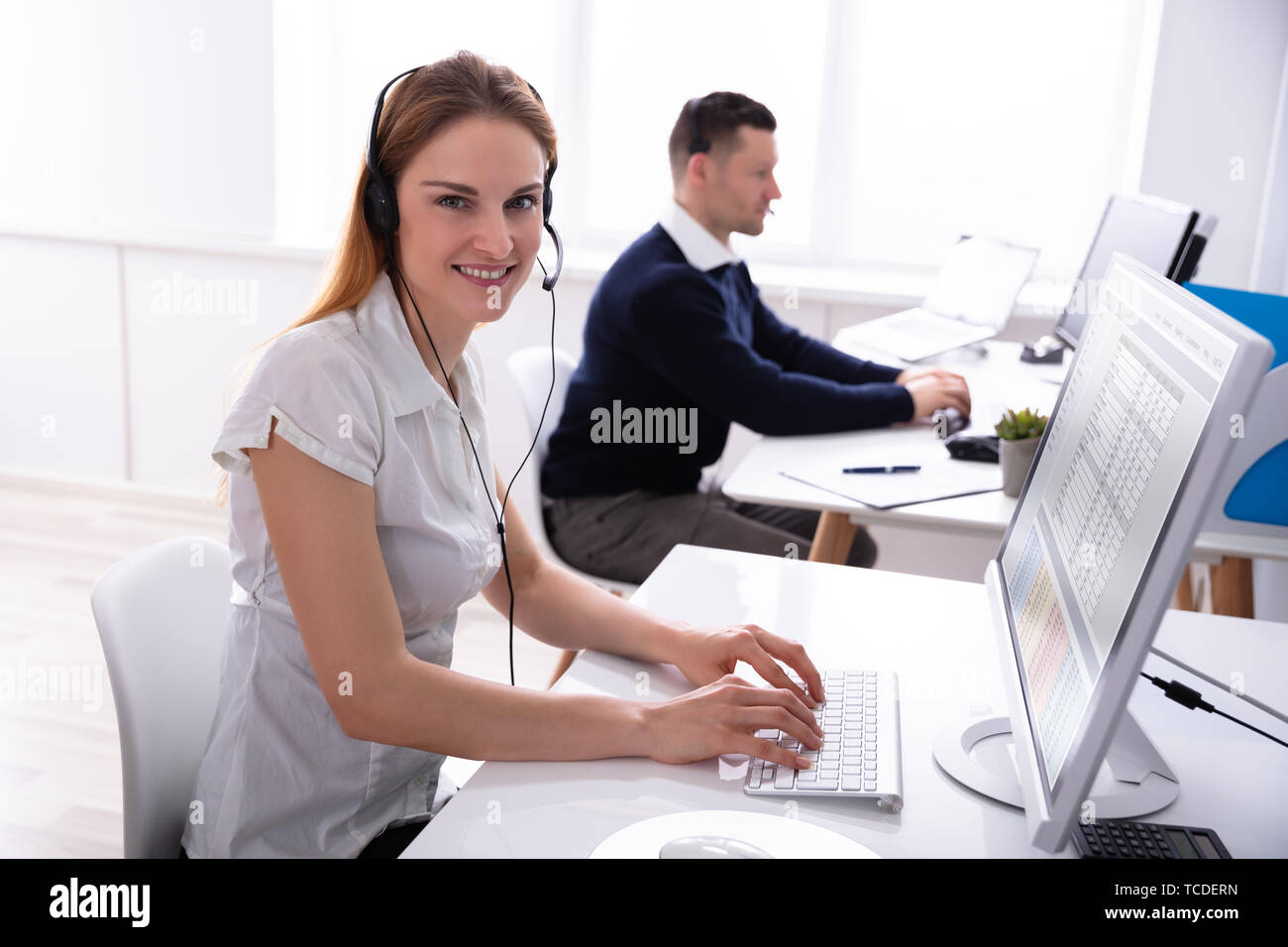 Portrait Of Young Smiling Businesswoman Using Computer Sitting On Chair Wearing Headset At Workplace Stock Photo