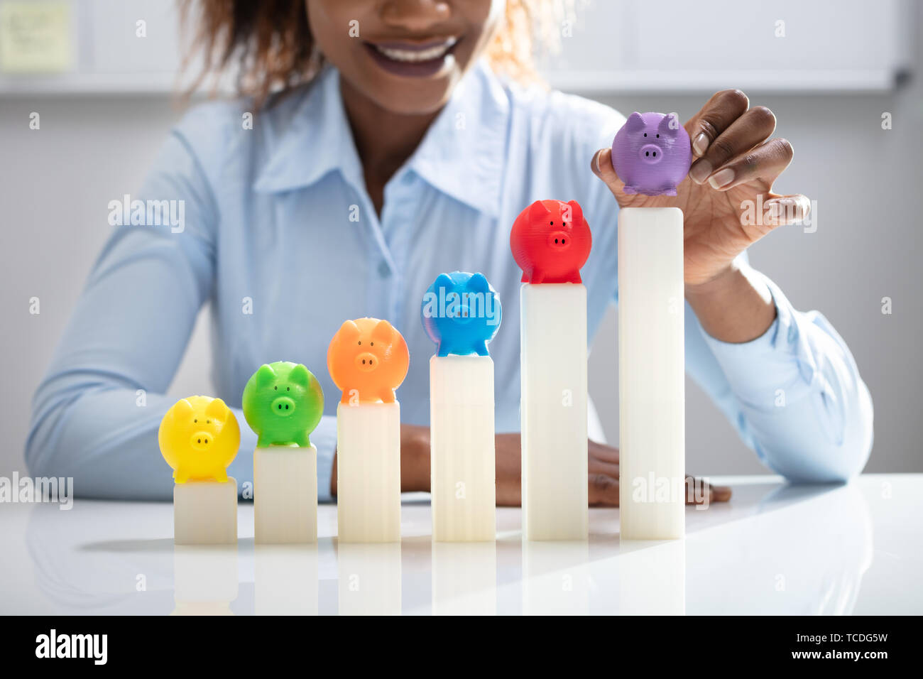 Businesswoman's Hand Placing Multi Colored Piggybank On Growing Graph In Office Stock Photo