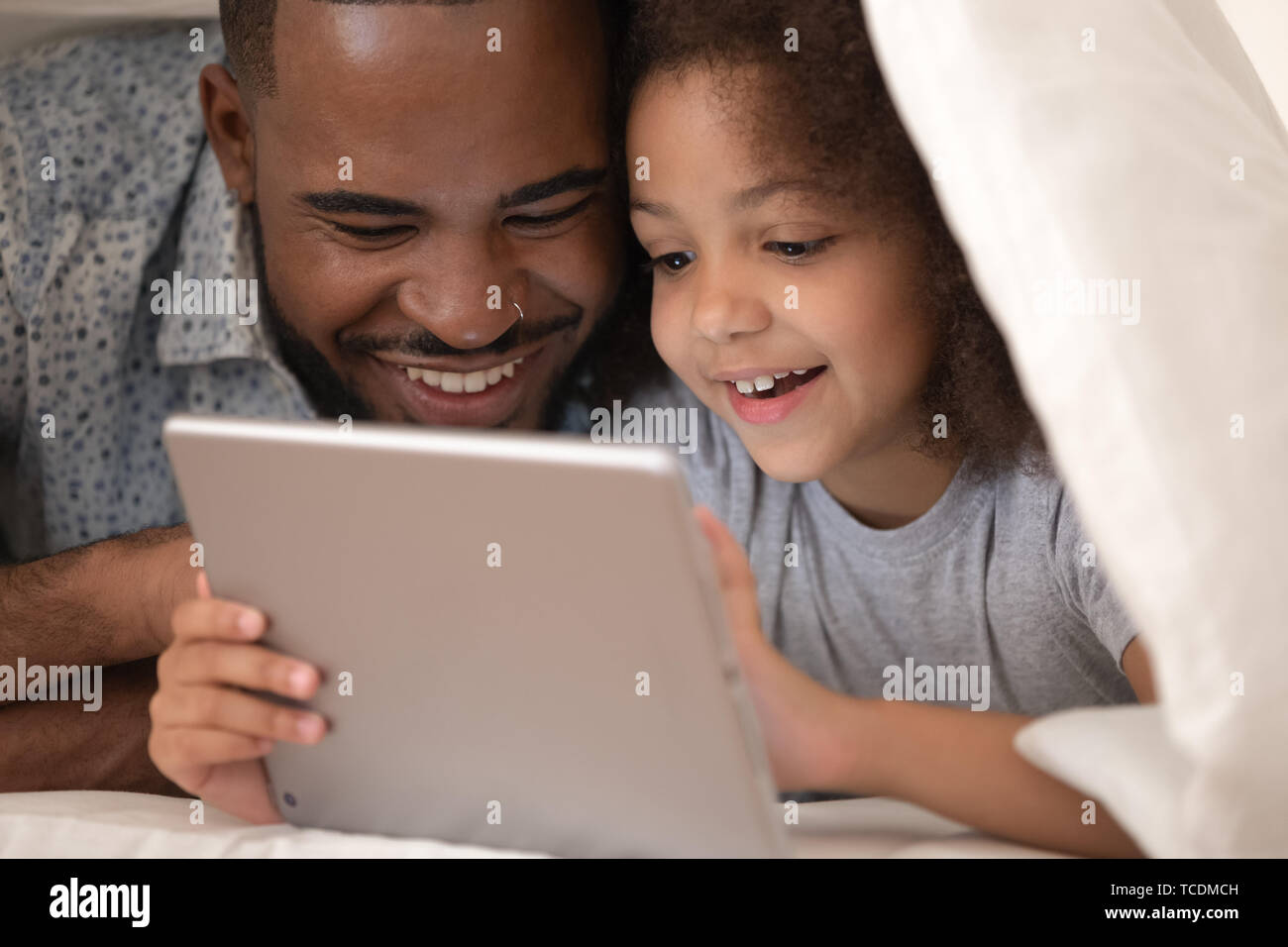 African father and daughter using tablet on bed under blanket Stock Photo