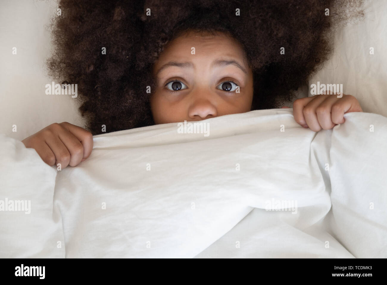 Scared african kid looking at camera covering blanket in bed Stock