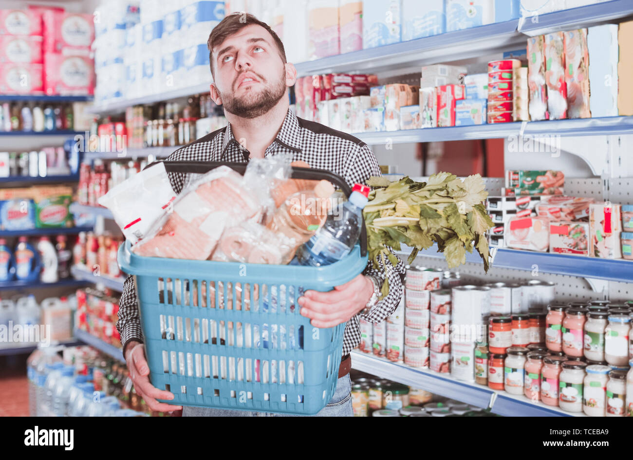 Tired young male with heavy shopping basket filled food products in supermarket Stock Photo