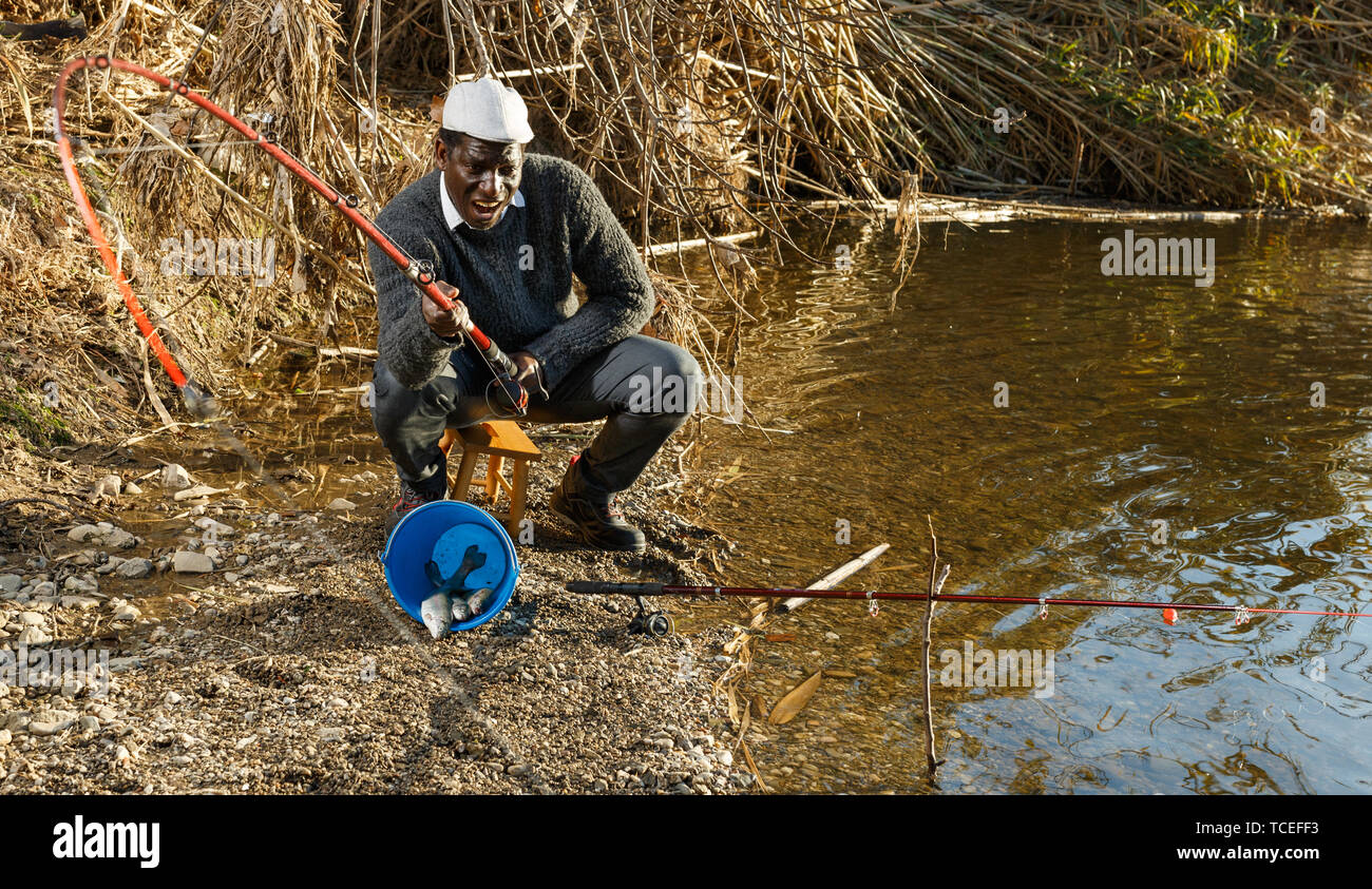 Mature African man sitting near river and pulling fish expressing emotions of dedication Stock Photo