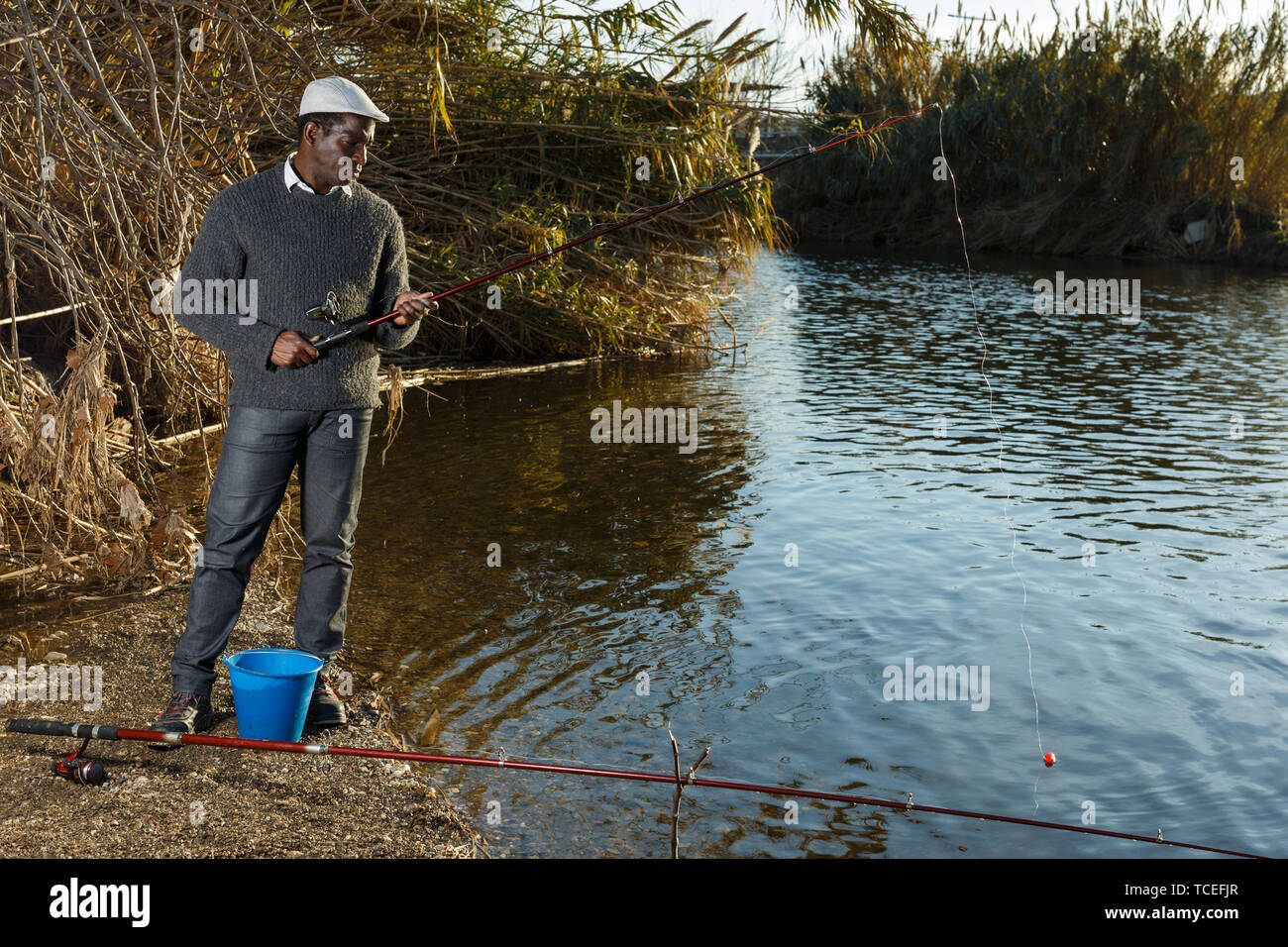 Mature African man standing near river and fishing with rods Stock Photo