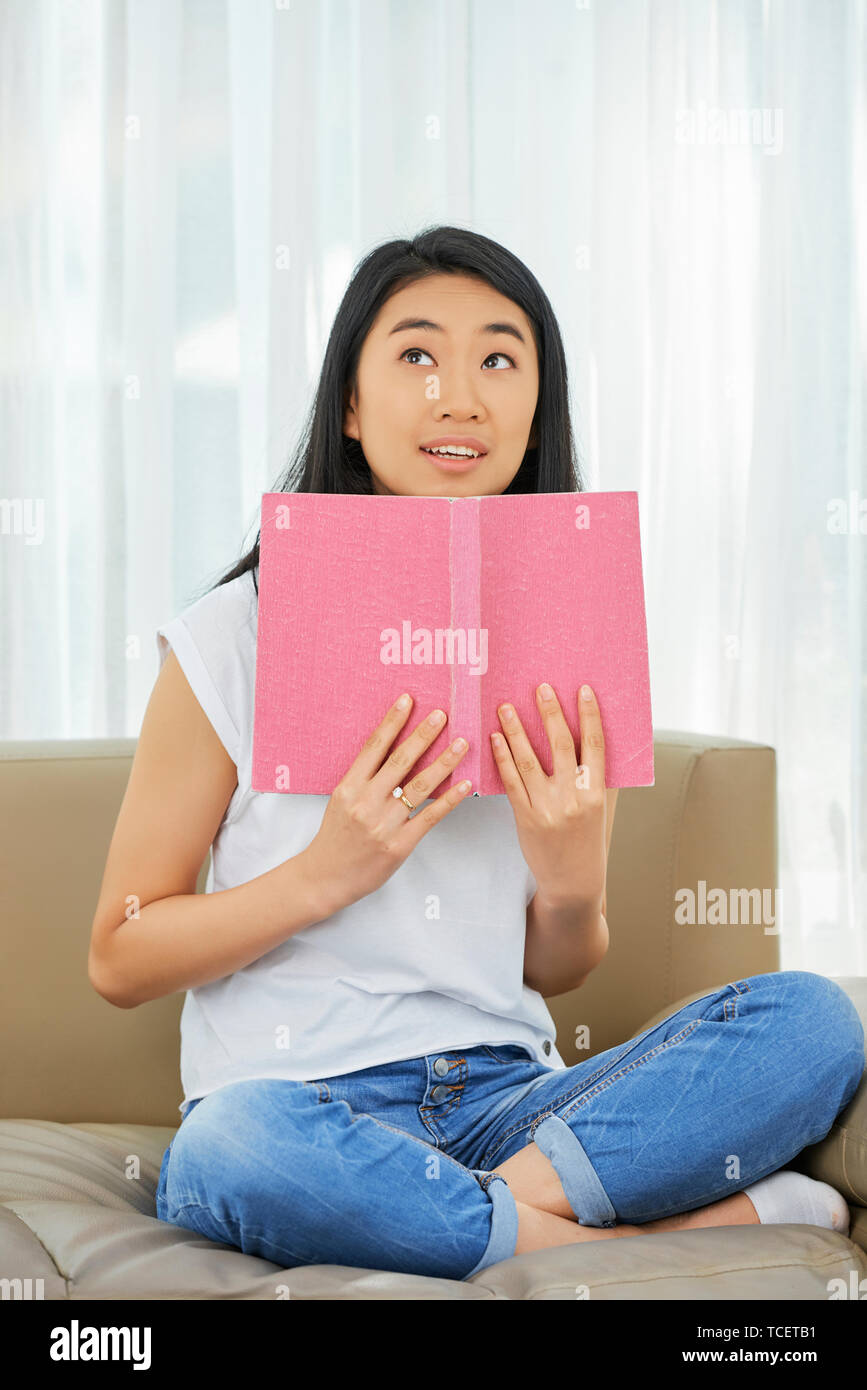 Portrait of positive Asian girl sitting on sofa with opened book and trying to memorize material for school Stock Photo