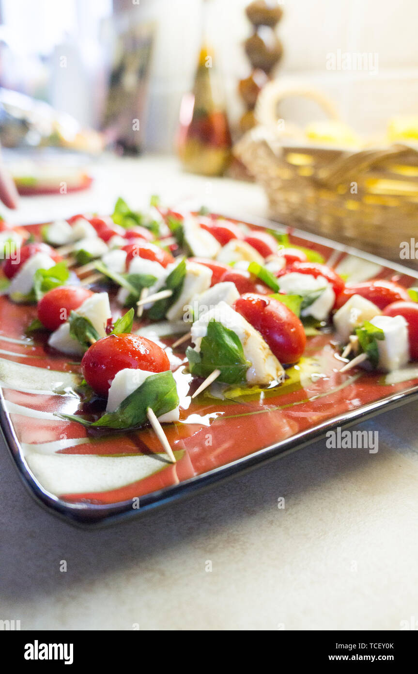 Cherry tomato with cheese and basilicas with olive oil on skewer lying in red dish with dark edge on kitchen in soft focus Stock Photo