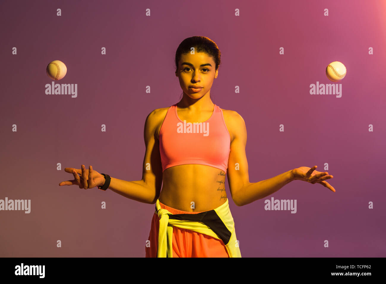 attractive african american girl in pink sports bra and shorts juggle with balls on purple background with gradient Stock Photo
