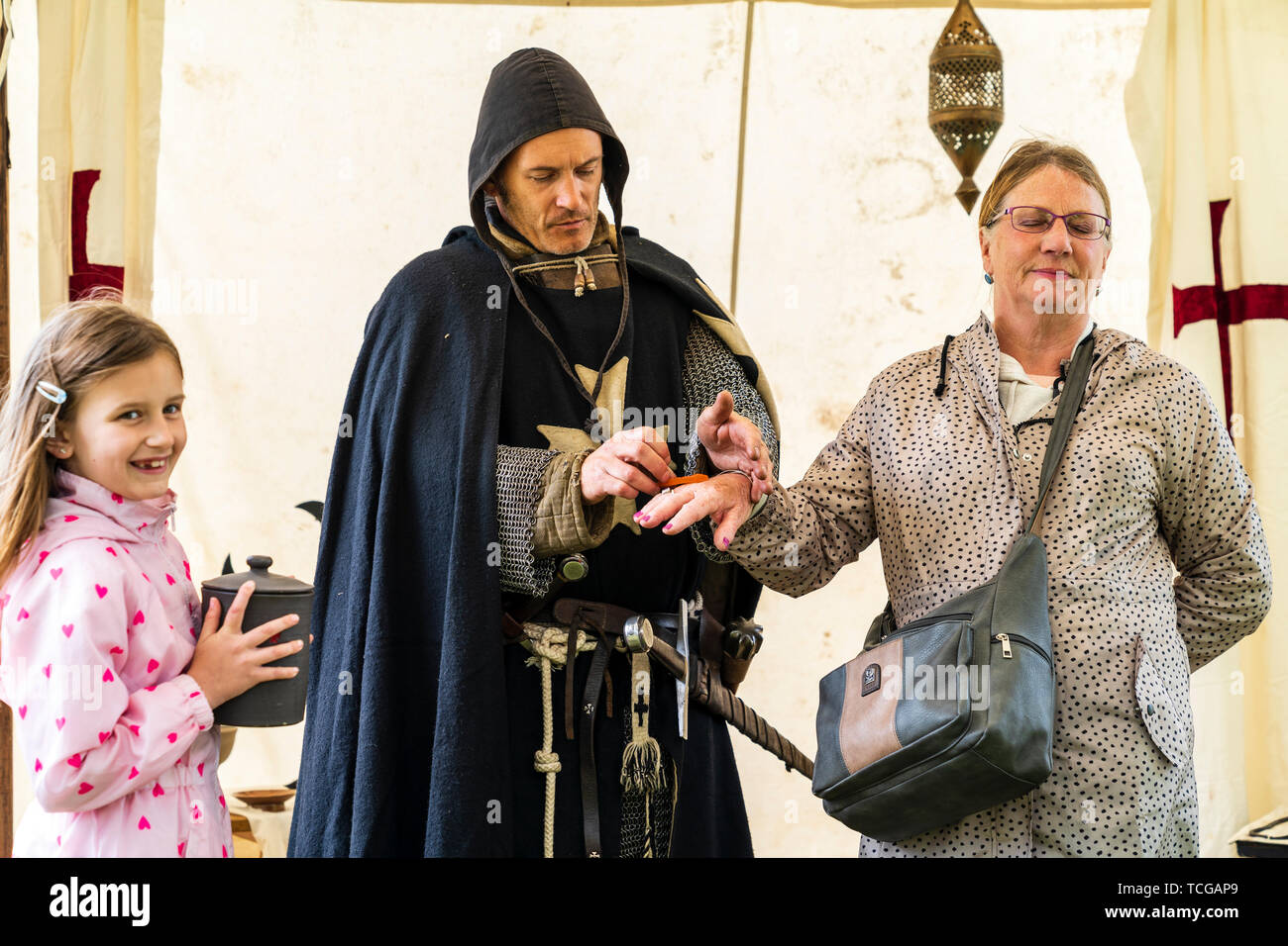 Medieval living history event. 12th century crusader in black cloak and chain mail, demonstrates the technic of 'leeching' on a mature woman member of the public with little girl, 7-8 year old, laughing next to him. Stock Photo