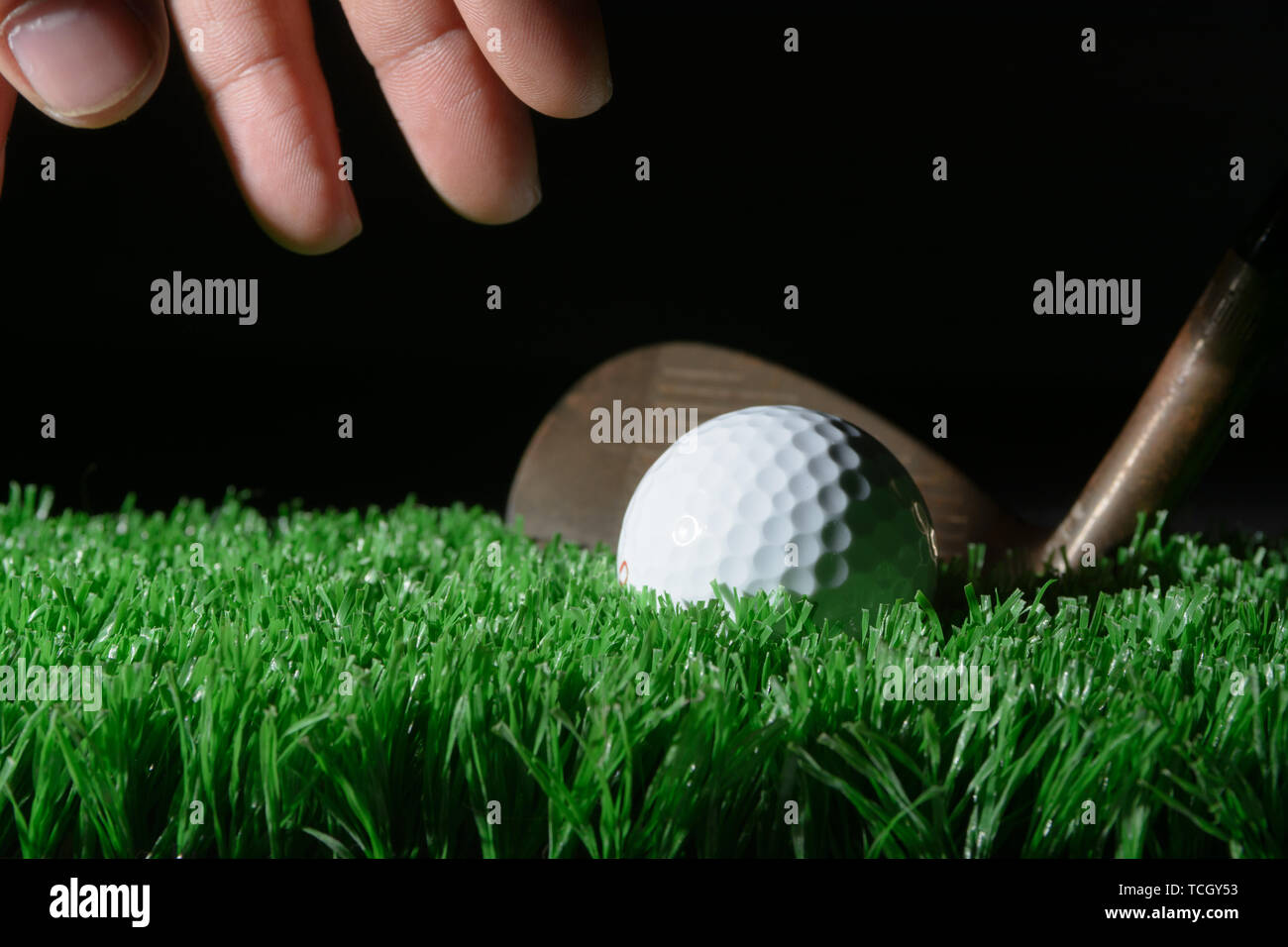 a hand grabs for the golf ball with a wedge sitting behind the golf ball on green grass. Stock Photo