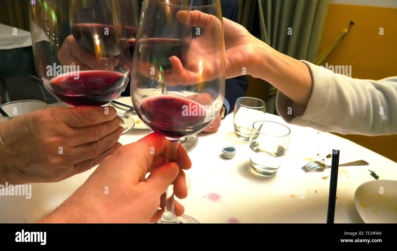 Close up of four hands with red wine glasses while doing cheers to celebrate in a restaurant, set table blurred on background. Stock Photo