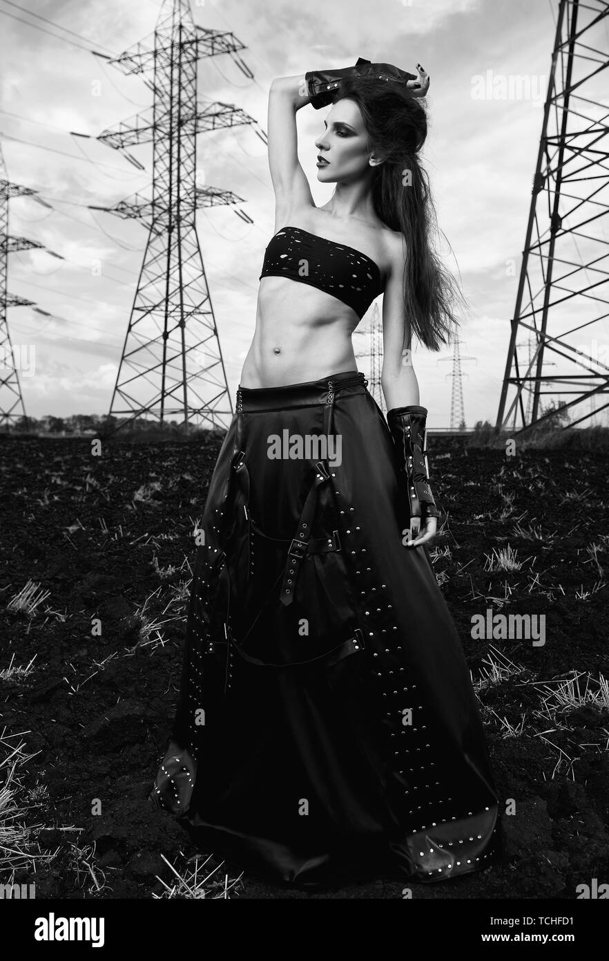 Informal fashion: the lovely slim young gothic girl dressed in black leather skirt and gloves. Outdoor portrait in field near power line towers. Black Stock Photo