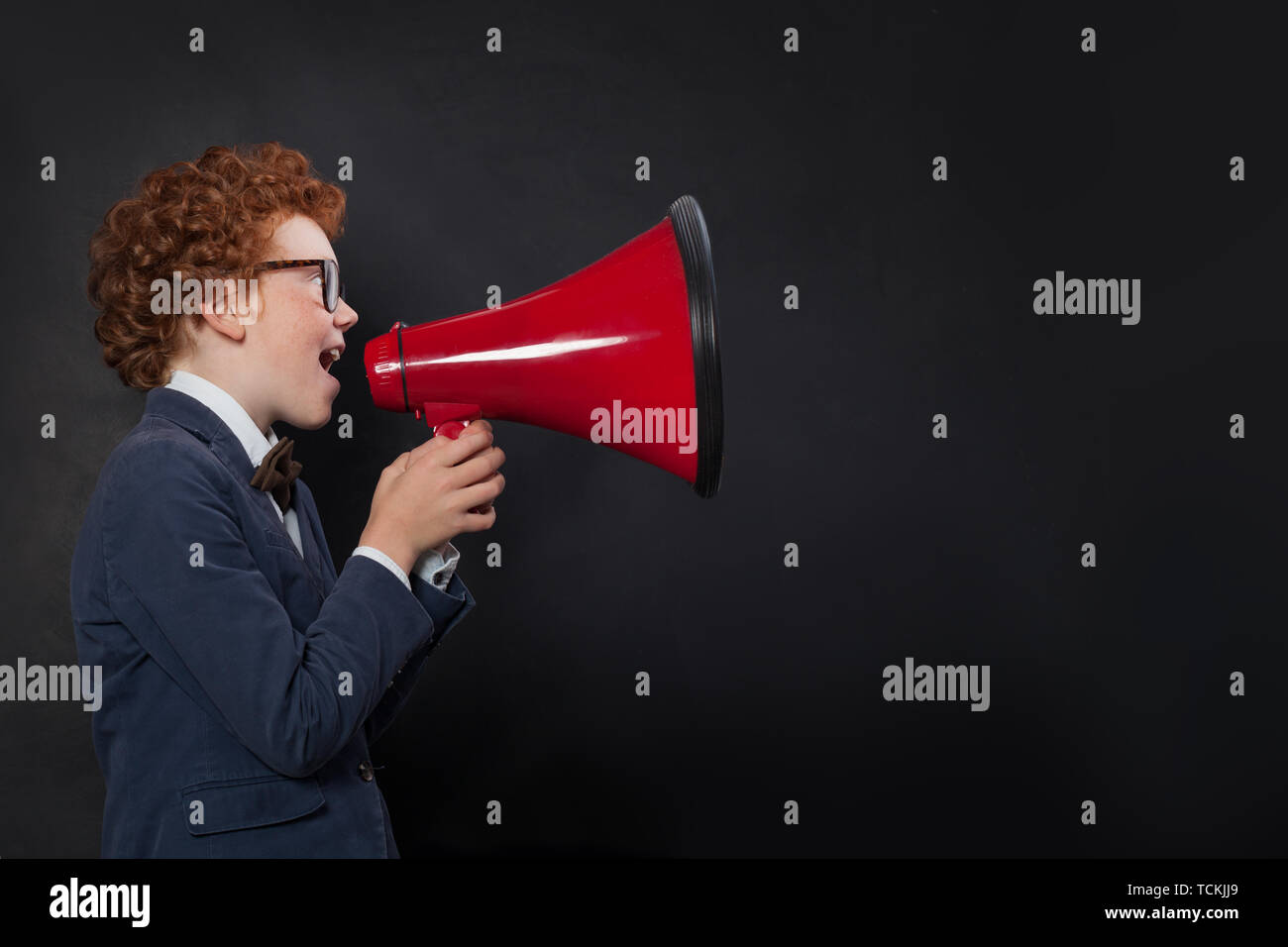 Cute child boy speaking through a megaphone against a blackboard with copy space. Kid with loudspeaker Stock Photo