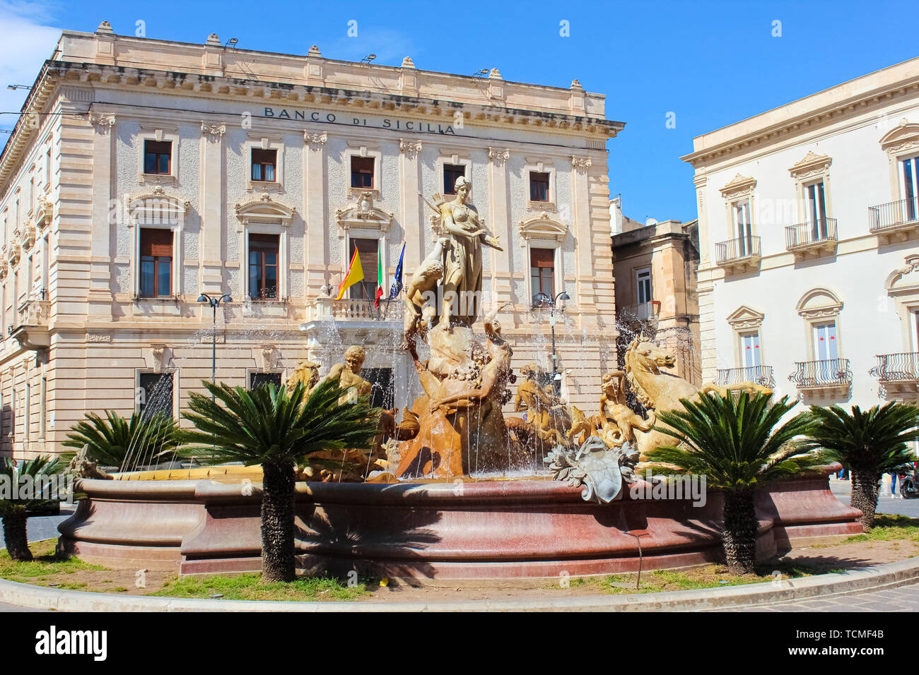 Syracuse, Sicily, Italy - Apr 10th 2019: Beautiful Fountain of Diana on the Archimedes Square in famous Ortigia Island. Building of a Sicilian bank in the background. Sunny day, blue sky. Stock Photo