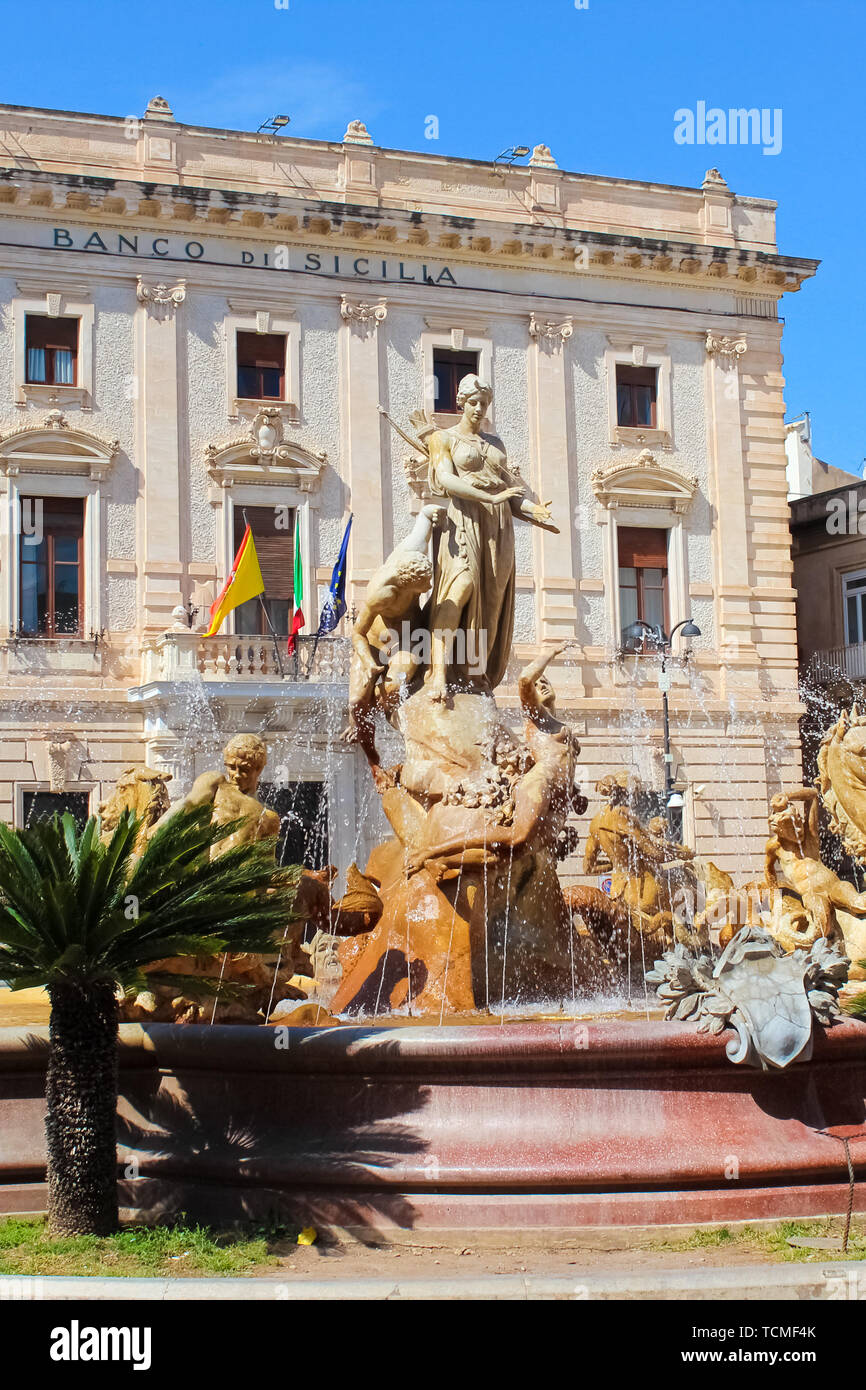Syracuse, Sicily, Italy - Apr 10th 2019: Amazing Fountain of Diana on the Archimedes Square in beautiful Ortigia Island. Building of a Sicilian bank in the background. Sunny day, blue sky. Stock Photo