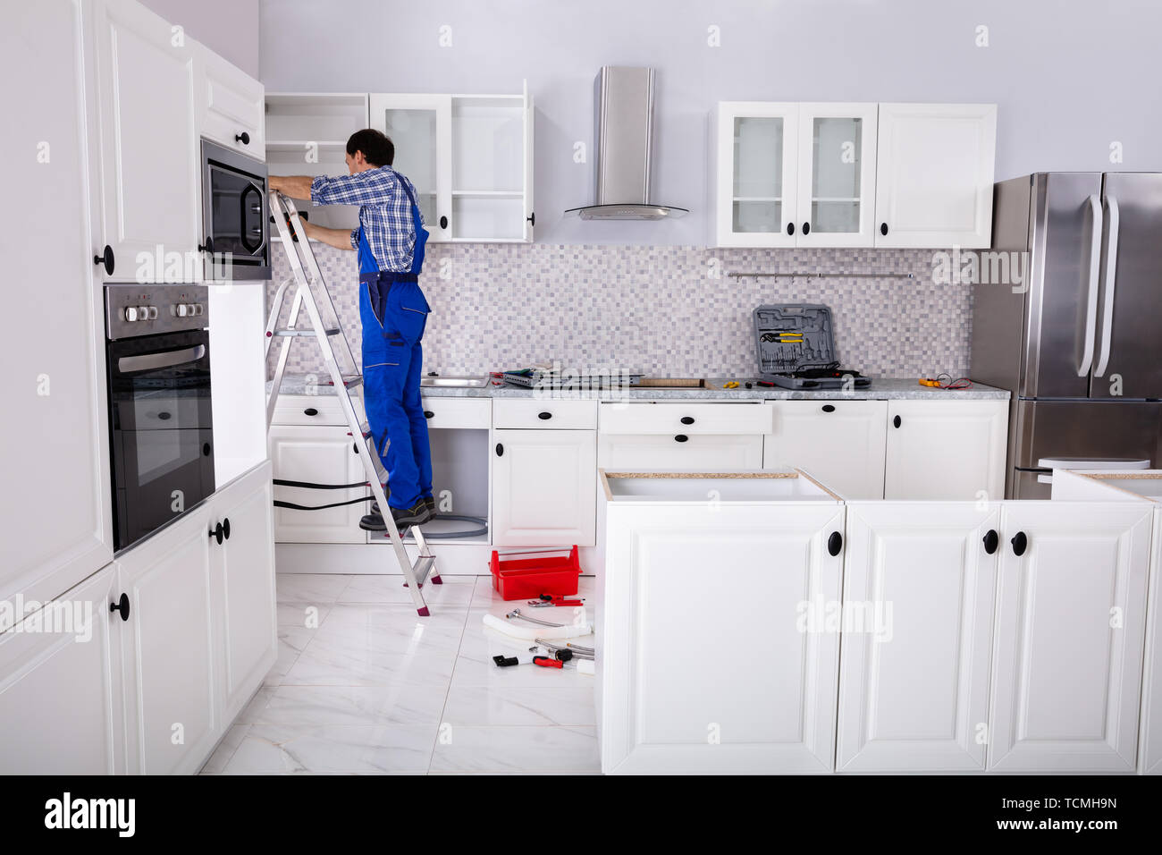 Rear View Of A Man Standing On Ladder Adjusting Cabinet Door Hinge