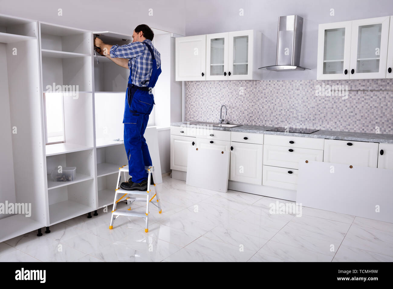 Rear View Of Serviceman Standing On Ladder Fixing Cabinet Door In