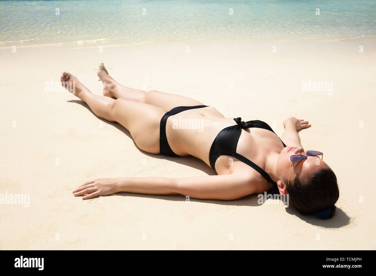 Young Woman In Black Bikini And Sunglasses Relaxing On Sandy Beach Near The Sea Stock Photo